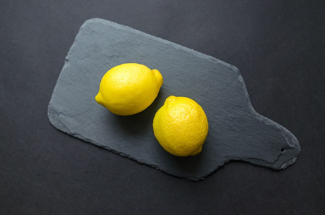 Photo prise du dessus de deux citrons jaunes posés sur une planche à découpée foncée.