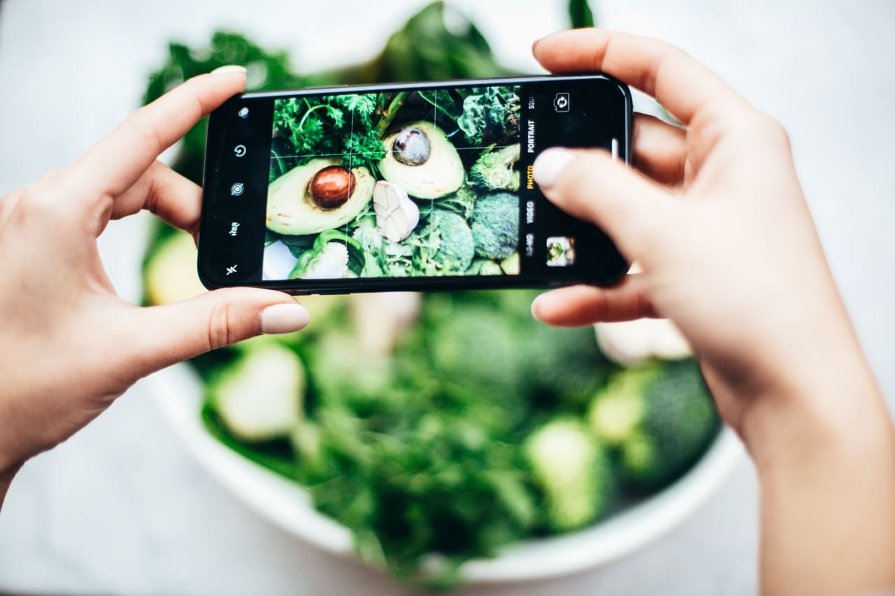 Une personne qui prend en photo avec un téléphone un plat composé d'avoat t de légumes verts. On voit l'écran du téléphone et donc la photo qu'est en train de prendre la personne.